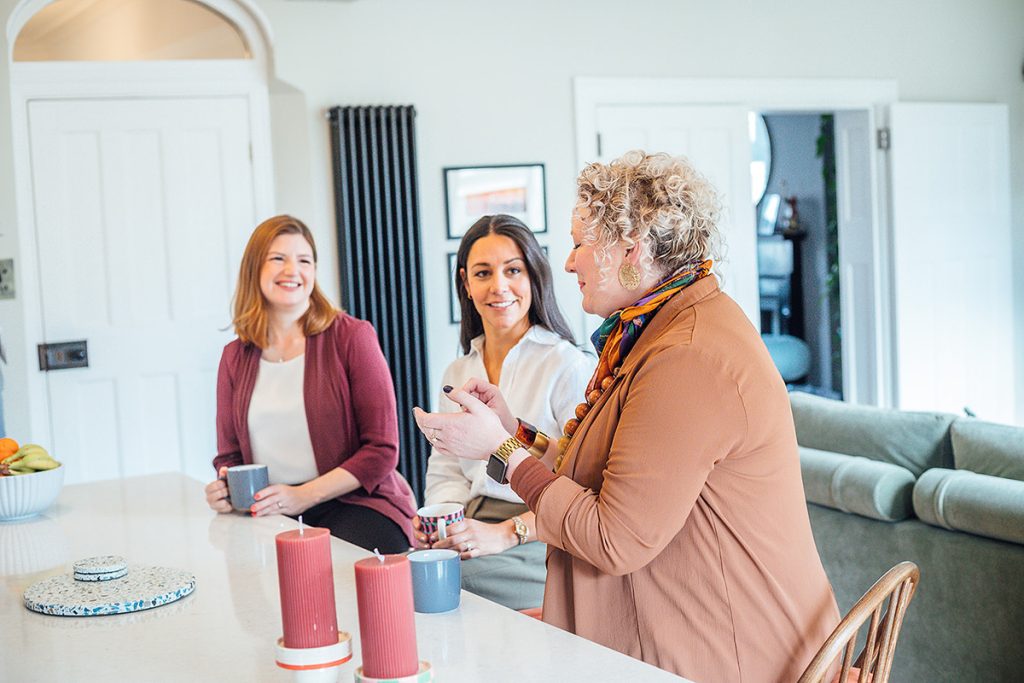 Three women chatting together in a bright, modern kitchen. One woman with short curly hair stands at a kitchen island, gesturing as she speaks, while the other two sit on stools holding mugs and smiling. Candles and a bowl of fruit sit on the counter, and a cosy living room is visible in the background, creating a warm, relaxed atmosphere.