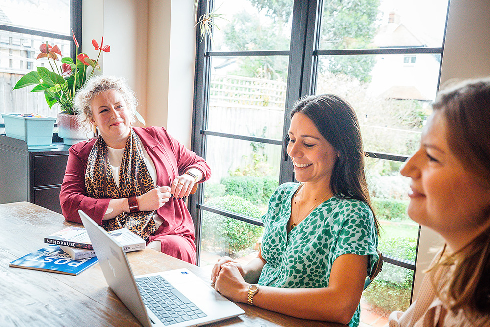 Three women sitting around a wooden table in a bright, modern room with large windows overlooking a garden. One woman in a green patterned dress is smiling while looking at a laptop in front of her. Another woman in a red suit with a patterned scarf sits opposite, watching attentively with a relaxed posture. A third woman is partially visible in the foreground. Books about menopause are on the table, suggesting a supportive discussion or consultation setting.