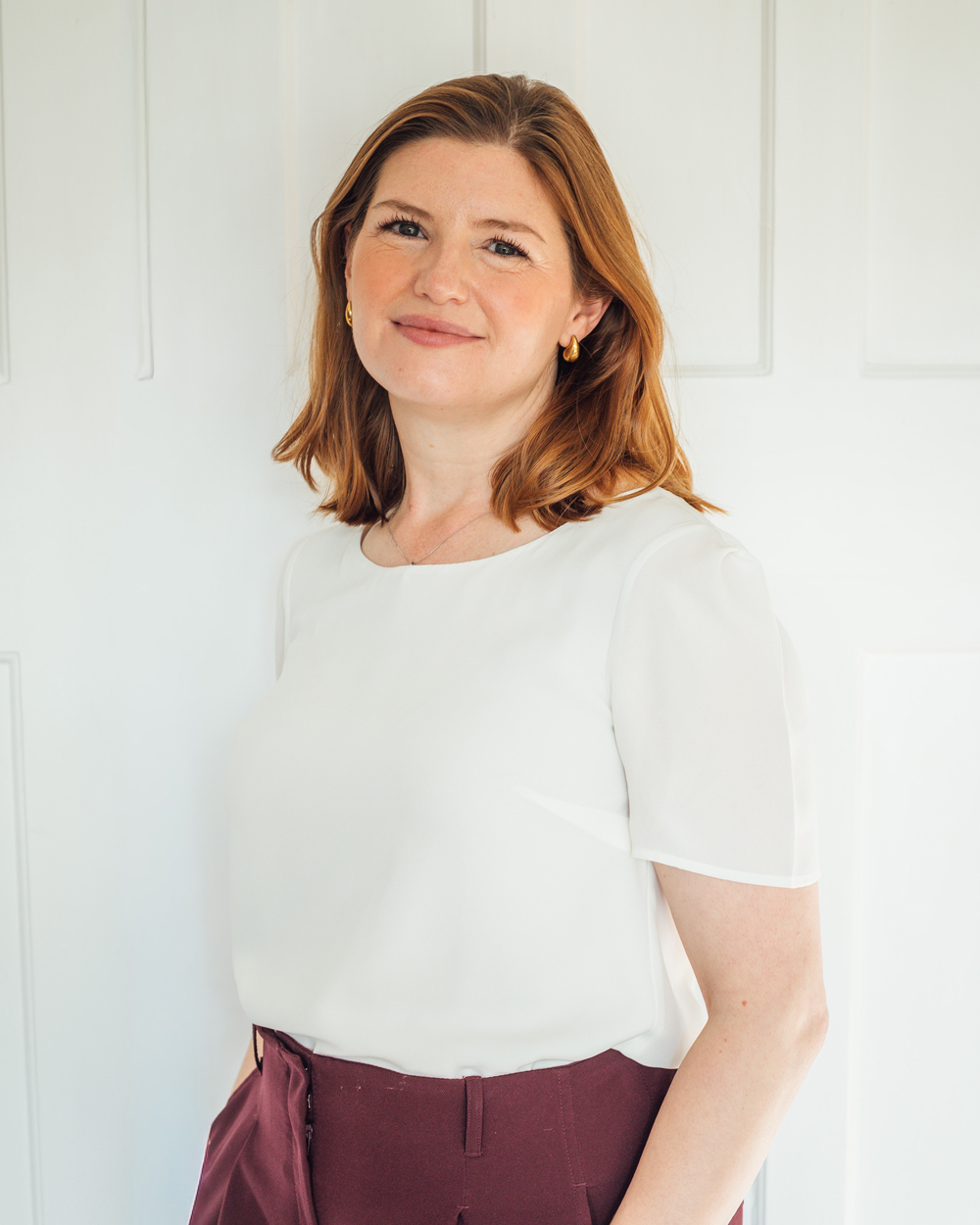 A woman with shoulder-length auburn hair stands against a light paneled wall, smiling softly at the camera. She wears a white blouse, burgundy trousers, and small gold hoop earrings, creating a clean, professional portrait.