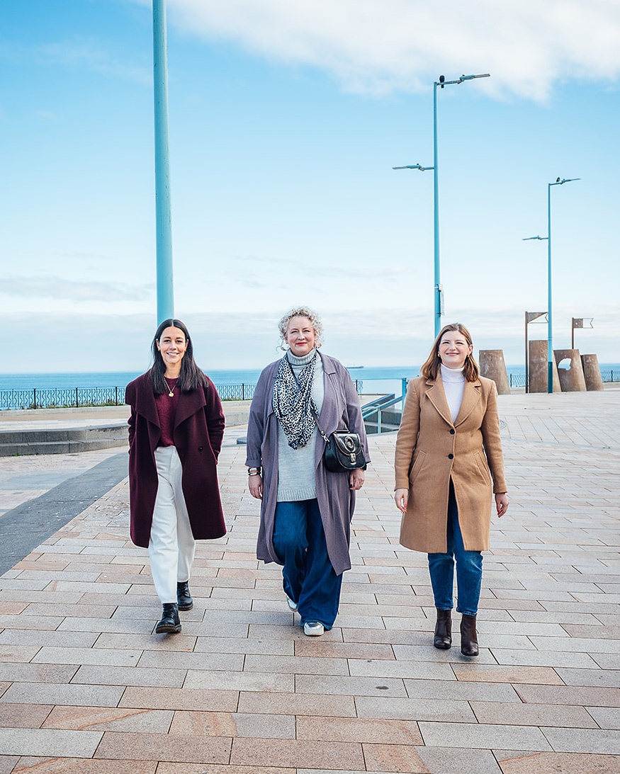 Three women walking side by side along a seafront promenade, wearing coats and smiling, with the sea and railings visible in the background.