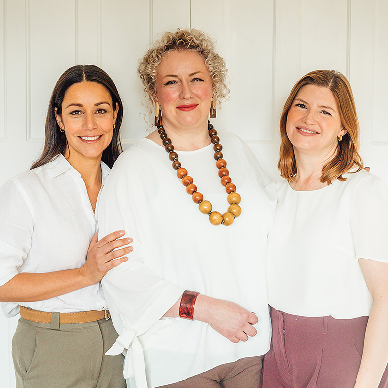 Three women stand together, smiling at the camera. The centre woman wears a white blouse with a large wooden necklace, while the others wear light tops and stand close beside her.