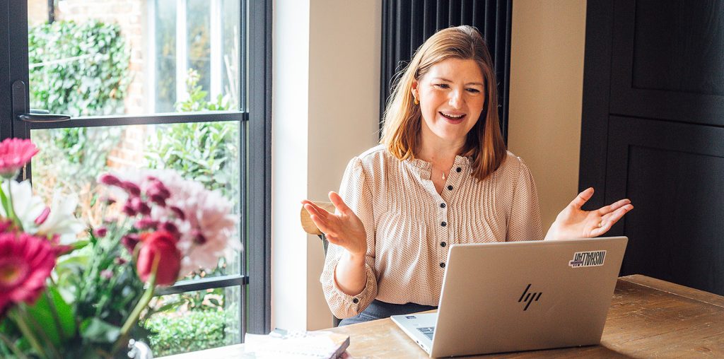 Ruth is smiling and gesturing while sitting at a wooden table with a laptop, beside a window with flowers in the foreground and natural light coming into the room.