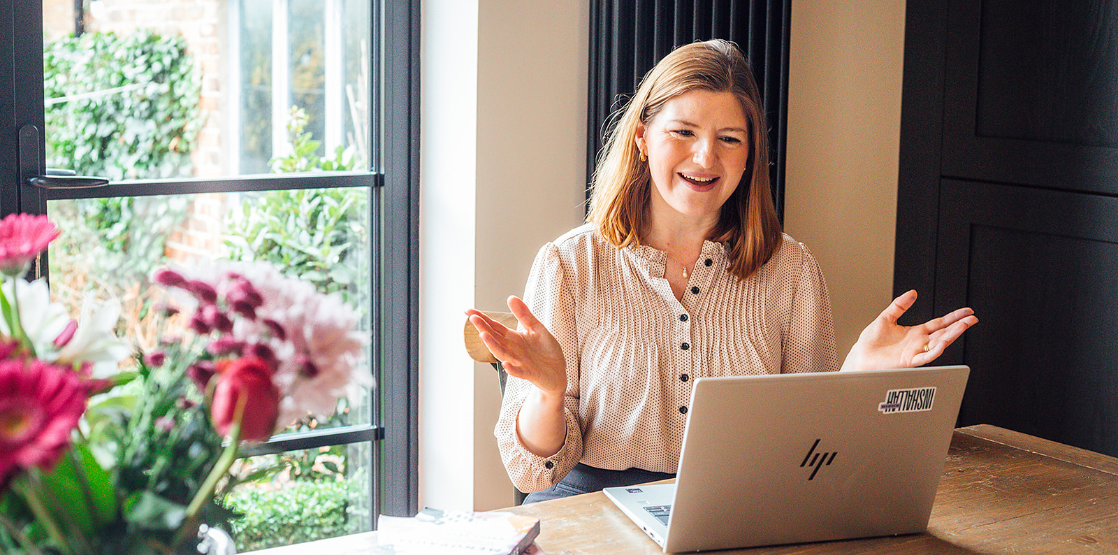 Ruth is smiling and gesturing while sitting at a wooden table with a laptop, beside a window with flowers in the foreground and natural light coming into the room.