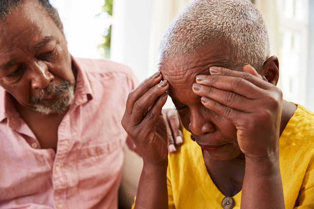 A man with a goatee and a pink button-down shirt comforts a woman with short, gray hair wearing a yellow shirt. The woman holds her face in her hands, appearing distressed.